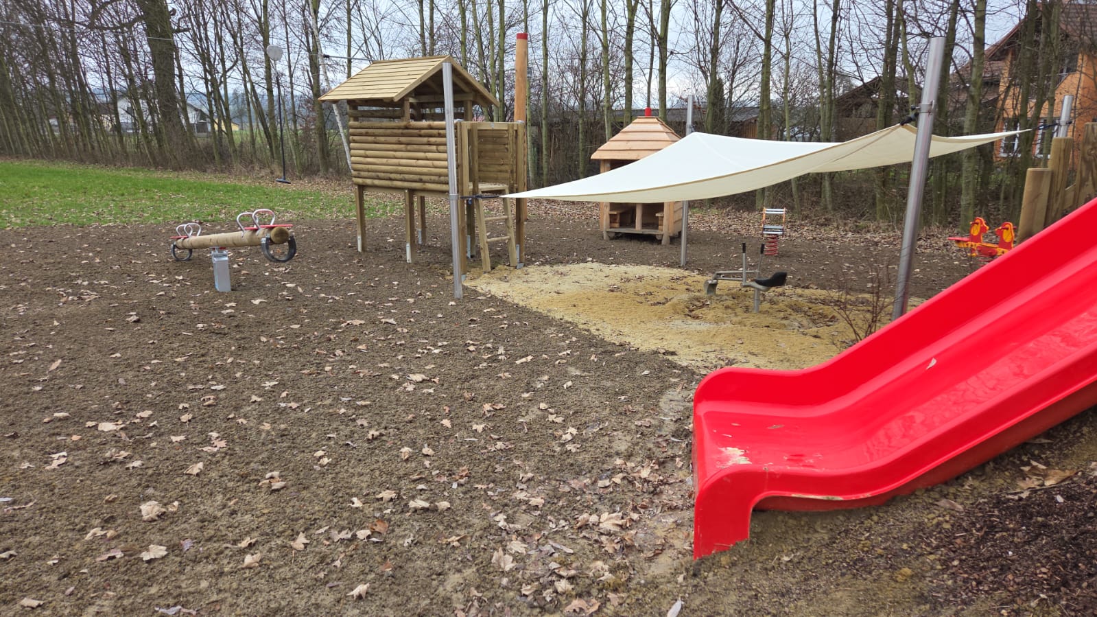Spielbereich mit roter Rutsche, Holzspielhaus und Sonnensegel in Hart und Münsteuer.