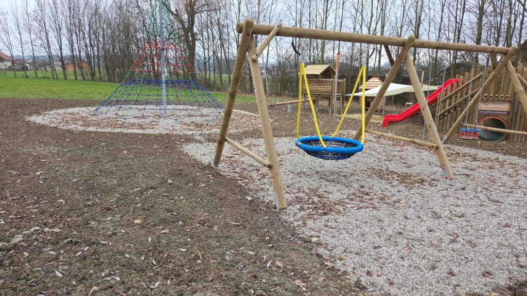 Detailansicht der blauen Nestschaukel und der roten Kletterpyramide auf dem Spielplatz.